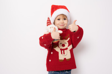 Little boy wearing santa hat holding santa claus toy with thumb up standing over white background.