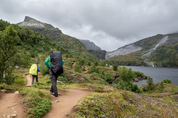 two hiker with backpacks on a path near a lake surrounded by high mountains and cloudy rainy sky on...