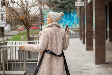 beautiful young woman with smart phone in hand wearing knitted hat and coat made of faux fur in...