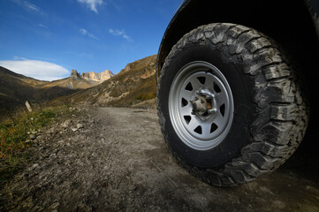 Fototapeta na wymiar Close-up A large off-road all-terrain car wheel stands on a rough road in the mountains against the backdrop of rocks. Off-road tours and off-road travel concept