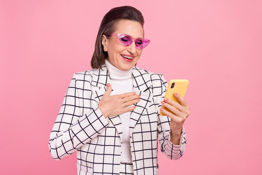 Portrait of cheerful lady in funky glasses hold hand chest see good news in smartphone isolated on pink color background
