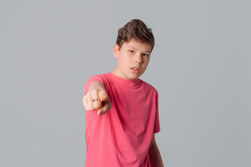 Portrait of frowning teenager boy and pointing finger to the camera and to you, wearing casual pink t shirt, standing over gray background