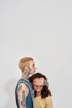 Two Diverse Young Women, Lesbian Couple Posing Together Isolated Over Gray Background. Woman Covering Her Girlfriend Eyes With Hand