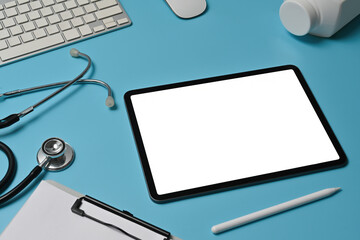 Top view image of a white blank screen digital tablet on the doctor table surrounded by a stethoscope, bottle pills, clipboard, wireless mouse, keyboard and stylus pen.