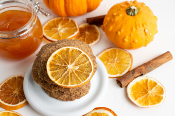 Close-up of stacked shortbread cookies with orange slices, cinnamon, pumpkins and jar of jam, selective focus, on white table, horizontal