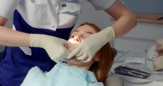 A female dentist in a medical uniform applies a retractor to a female patient and with an assistant performs a professional dental cleaning procedure in a modern dental office. 