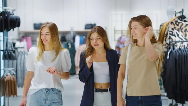 Stylish young women walking in clothes store together, talking and pointing at apparels. Cheerful friends shopping in mall. Concept of consumerism
