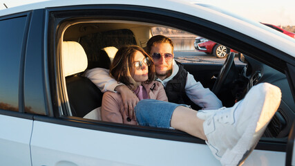 Happy young carefree tourists travelers couple sitting in car at sunset in countryside. People, tourism, lifestyle concept