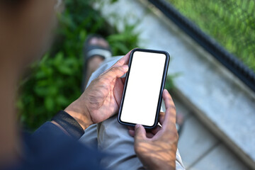 Top view image of a man's hand holding a white blank screen smartphone while sitting next to the wire mesh fence.