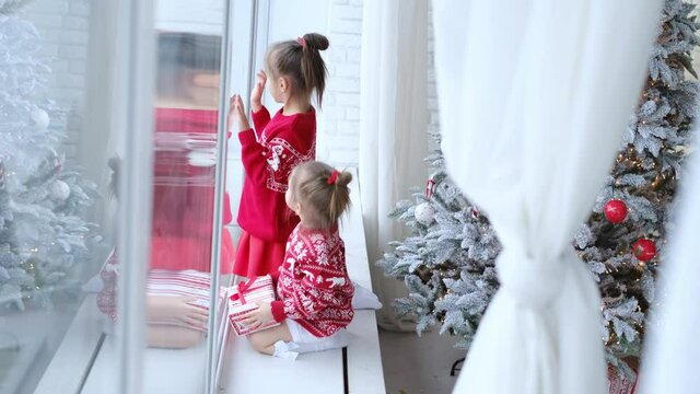 Girls in warm New Year sweaters sit on the windowsill, draw with their finger on the glass and watch it snow outside the window. Children are waiting for Christmas miracles. xmas