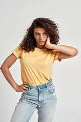 Confident young female model with dark curly hair in casual wear looking at camera while posing isolated over gray background