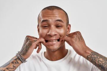 Portrait of funny mixed race tattooed guy looking at camera, forcing smile with fingers in his...
