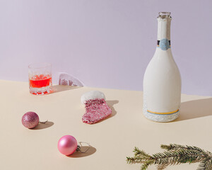 Minimal New Year composition with Christmas decoration, champagne bottle, and a glass of drink on pastel purple and yellow background.