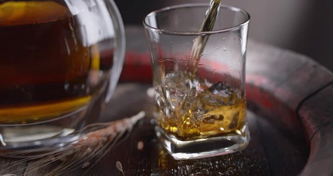 Jet of whiskey is poured into a glass with ice cubes, standing on a vintage whiskey barrel. A decanter with a drink stands nearby, a dark brown background.