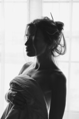 Black and white portrait of blond young beautiful woman having fun hiding under white sheet standing on light window. Beautiful girl stands at the window covered with a blanket