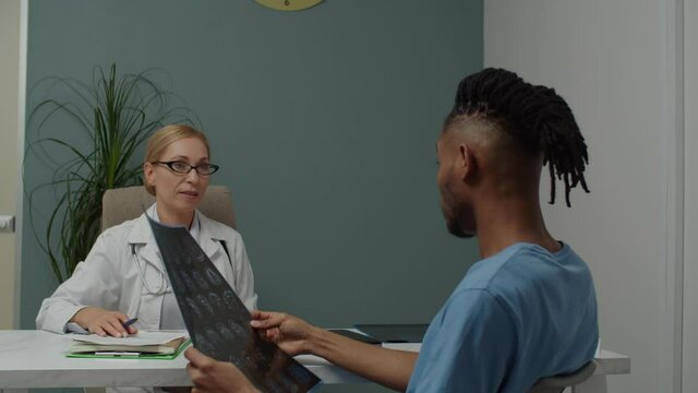 Young adult male patient in casual clothing looking at MRI image, questioning female doctor about result, diagnosis indoors while having physician's appointment in hospital or clinic