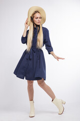 High fashion photo of a beautiful elegant young woman in a pretty blue dress, hat, boots posing over white, soft gray background. Studio Shot, portrait. Blonde