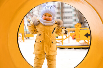 A baby aged 12-17 months , looks into a yellow pipe on the playground in winter