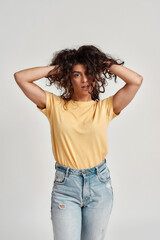 Playful young woman with dark curly hair in casual wear looking at camera, adjusting her hair while standing isolated over gray background