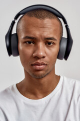 Closeup portrait of serious mixed race guy wearing headphones looking at camera, listening to music or podcast while posing isolated over white background