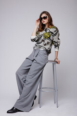 Fashion photo of a beautiful elegant young woman in a pretty gray pants, trousers, blouse, bicolor accessories, boots posing over white, soft gray background. Brown hair. Studio Shot. 