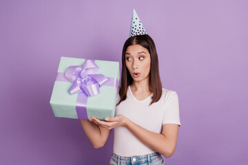 Portrait of young girl wearing paper cap holding in hands giftbox amazed surprised shock isolated over vivid violet color background