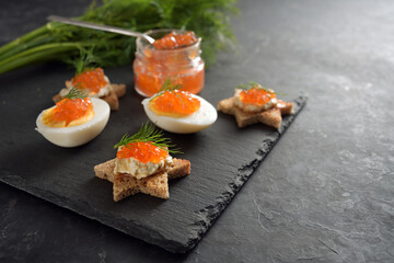 Red caviar on halved eggs and on toasted canapes in star shape with dill, preparation for a festive Christmas buffet on a dark slate plate, large copy space