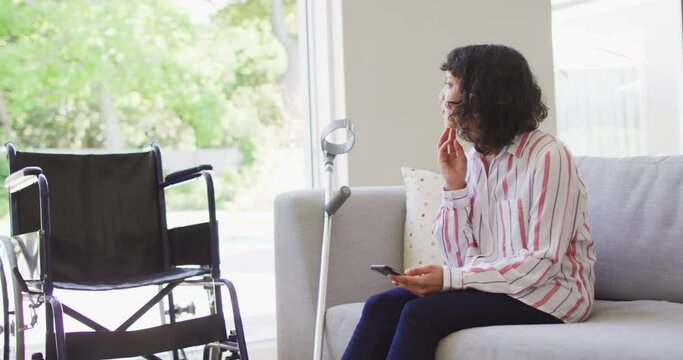 Thoughtful biracial disabled woman using smartphone in living room, crutch and wheelchair by couch