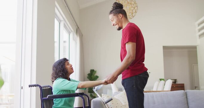 Happy biracial woman in wheelchair and smiling male partner holding hands and talking in living room