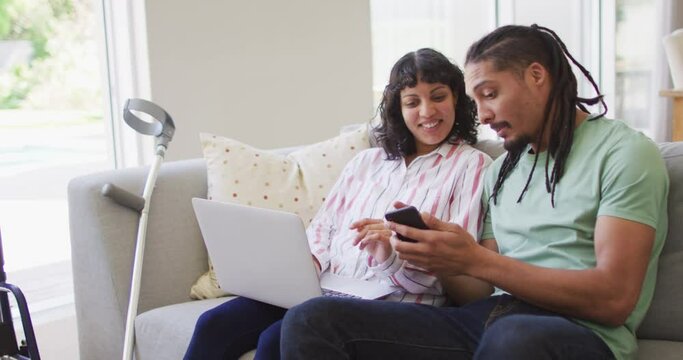 Happy biracial couple in living room using laptop and smartphone, with crutch leaning on couch