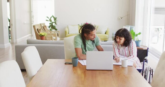 Happy biracial woman in wheelchair and male partner using laptop and talking in living room