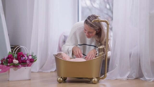 Wide shot happy mother washing baby daughter in golden bath in living room at home. Portrait of positive beautiful Caucasian woman taking care of newborn infant baby girl. Slow motion