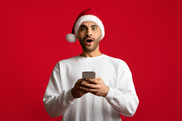 Amazing Offer. Portrait Of Surprised Arab Man In Santa Hat Holding Smartphone