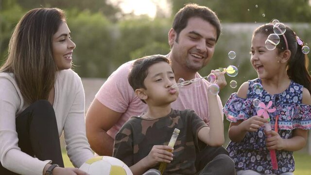 Happy Indian family including mother, father, and two kids are blowing soap bubbles and enjoying the summer holidays in the park. Smiling parents and children are spending leisure time together.