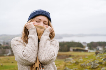 Young woman in knitted jaket holding scarf with hands over nose. Nature in background. Cloudy day in Norway. Closed eyes.