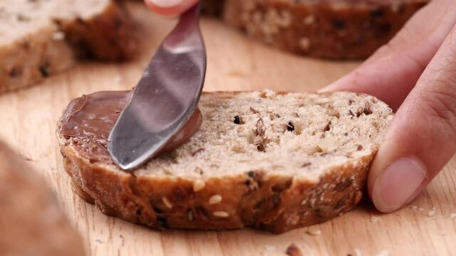 Women holding sliced multigrain bread on wooden table and using butter knife spreading tasty chocolate cream on fresh bread with seed.