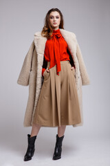 High fashion photo of a beautiful elegant young woman in a pretty beige coat, long skirt, red bow...