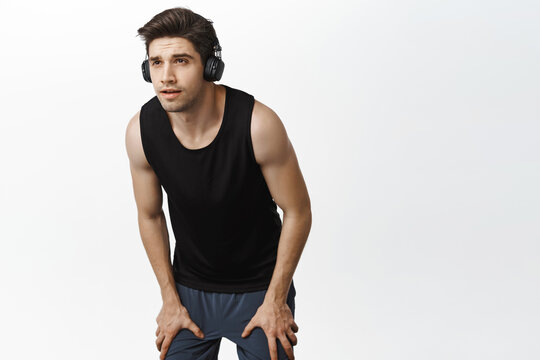 Strong and healthy male runner workout in wireless headphones, panting and looking ahead while jogging, taking break, standing over white background