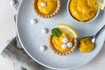 Homemade lemon curd in tartlets with fresh lemon slices, mini meringue and mint on ceramic plate, light concrete background. Top view.