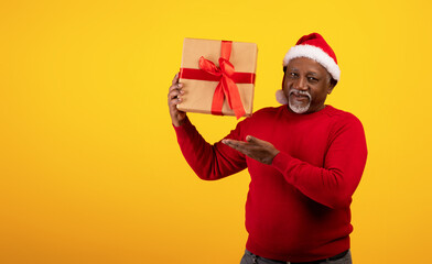Christmas portrait of joyful senior black man in Santa hat holding Xmas gift over orange background, empty space