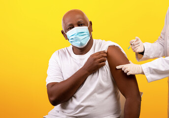 Doctor giving coronavirus vaccine shot to senior African American male patient on orange studio...