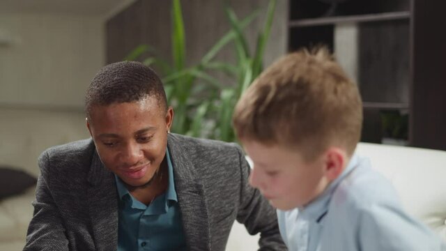 Young African-American English teacher nods head to cheer up junior schoolboy done task right at private tuition with native speaker in room slow motion