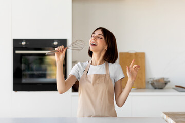 Fototapeta na wymiar Playful woman in apron singing a song, pretending whisk to be a microphone, having fun in modern kitchen interior
