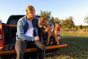 Two best friends, young man and canine, nurturing their friendship and building mutual trust while sitting on the back of a pickup truck - obrazy, fototapety, plakaty