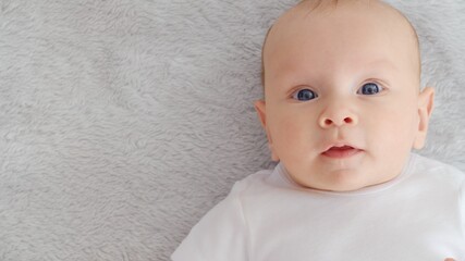 Child's face of a Happy newborn baby looking to camera. Healthy newborn baby in a white t-shirt with blue eyes. Face of a Beautiful active tiny child. Cute white Infant boy, top view.