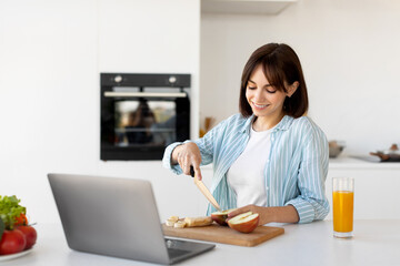 Millennial lady cooking fruits salad, cutting apple and banana, watching food blog videos on laptop computer, free space