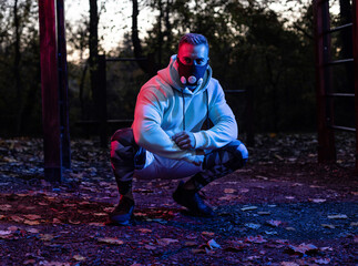 Strong good looking young man with workout fitness mask in squatting position in the park taking a break from workout as dusk sets, hard street workout