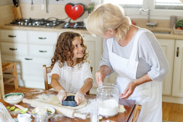 Adorable curly little girl with her grandmother cooking homemade cookies together at kitchen table.