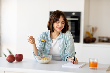 Happy young woman enjoying breakfast, eating cereals with orange juice and making notes in notepad,...