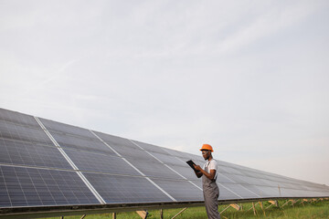 African american industrial worker in uniform and safety helmet standing among field with solar...